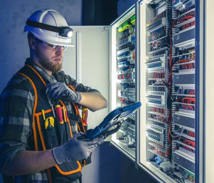 Electrician focused on work in switchboard with fuses, using tablet. Young adult electrical engineer in special clothes with flashlight on helmet in dark room with emergency lights in background.